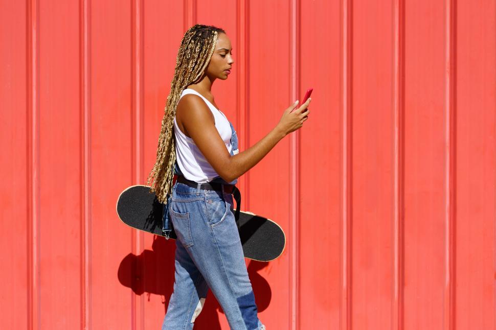 Free Image of Black woman with coloured braids, consulting her smartphone with her feet resting on a skateboard. 