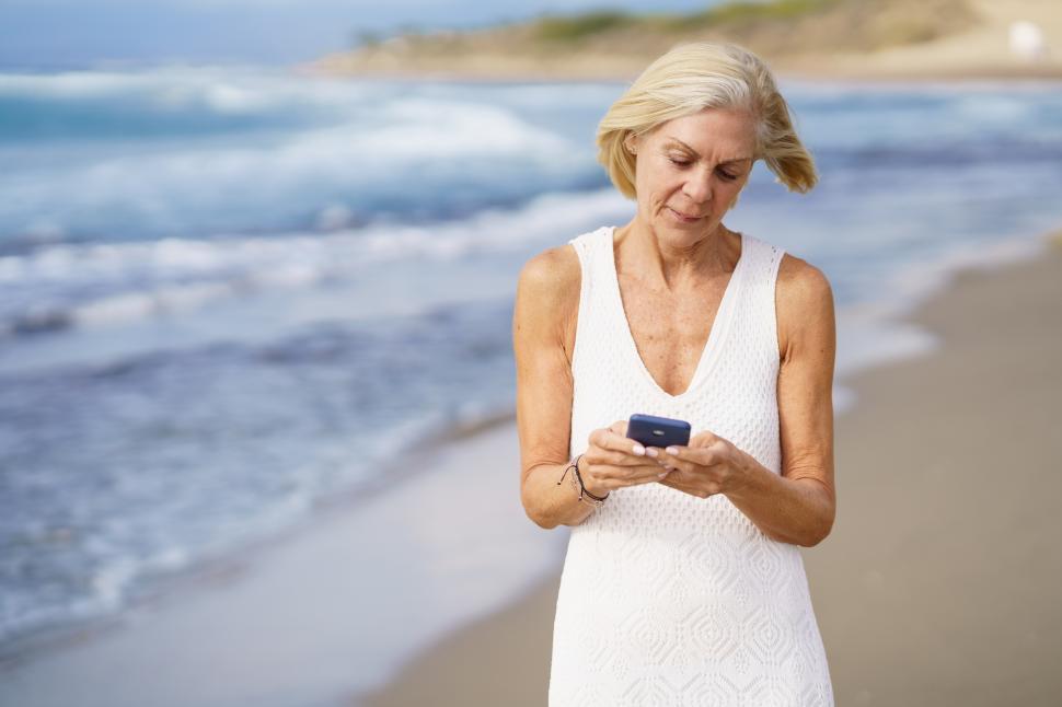 Free Image of Senior woman walking on the beach using a smartphone. 