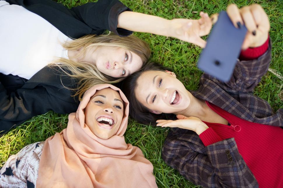 Free Image of Multiracial girlfriends laughing while lying on lawn and taking selfie 