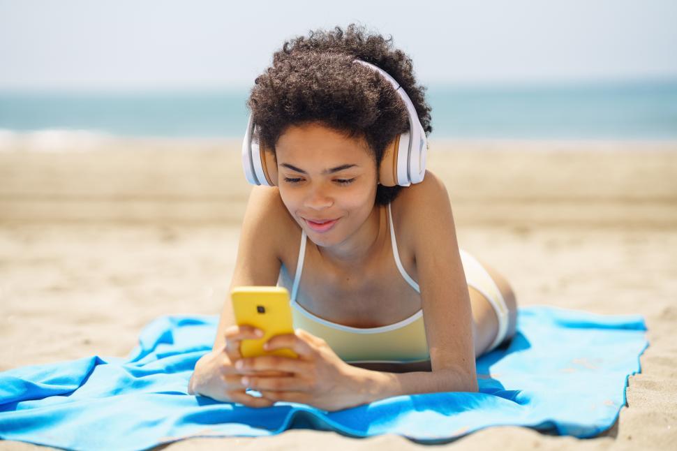 Free Image of African American woman lying on a towel on the sand on the beach using her smartphone 