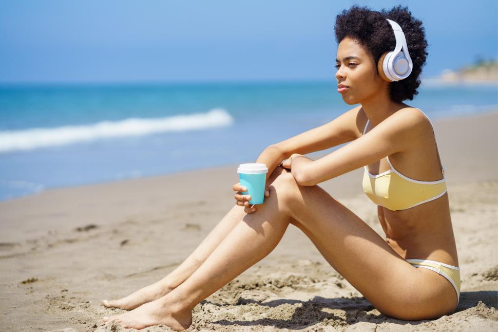 Free Image of Black woman in headphones drinking takeaway beverage while relaxing on beach 