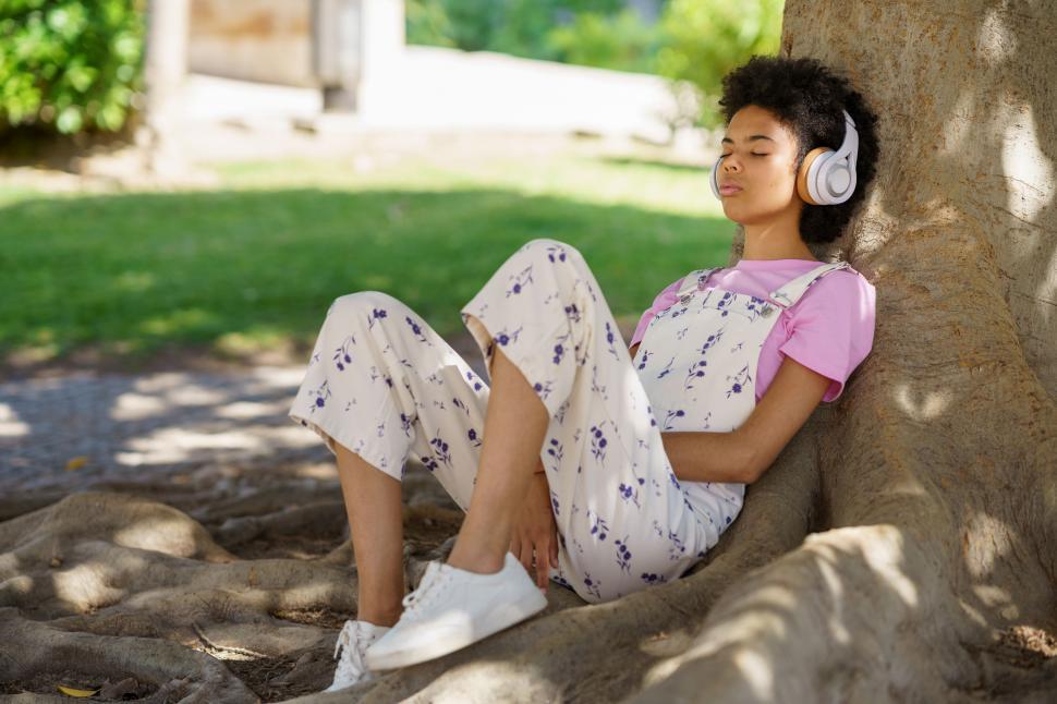 Free Image of Black woman sitting in the shade of a tree relaxing and listening to music with headphones. 