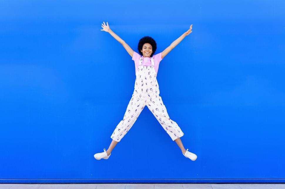 Free Image of African American female jumping with raised arms against a blue urban wall. 
