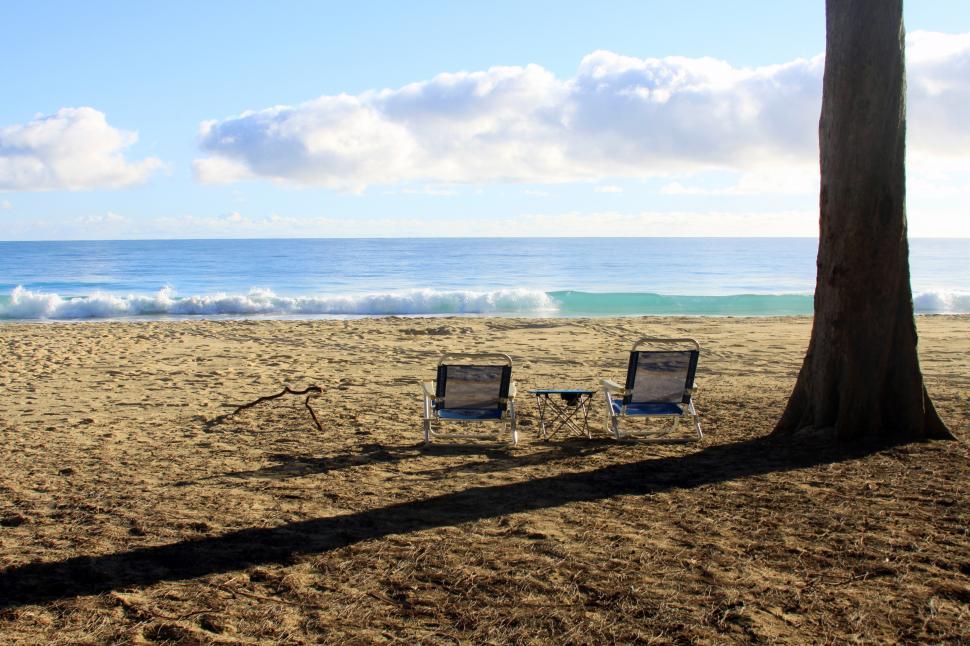 Free Image of Chairs on a beach with a tree and water in the background 