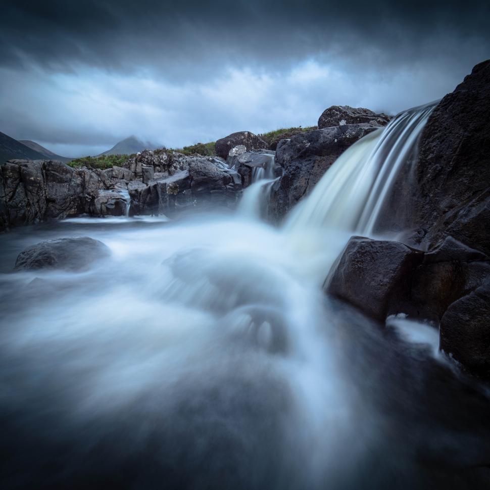 Free Image of A waterfall over rocks with mountains in the background 