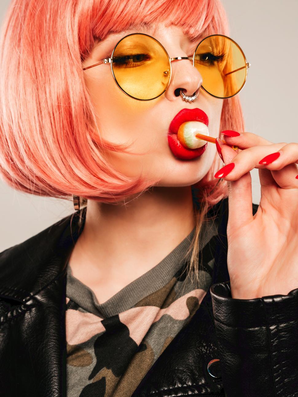 Free Image of A woman with pink hair and yellow sunglasses eating a lollipop 