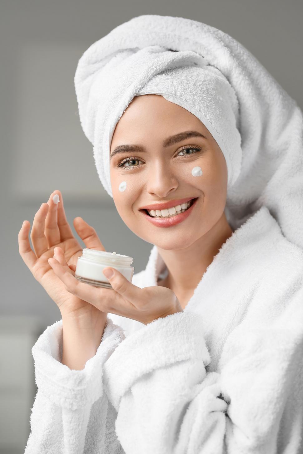 Free Image of A woman wearing a white robe and a towel on her head holding a jar of cream 