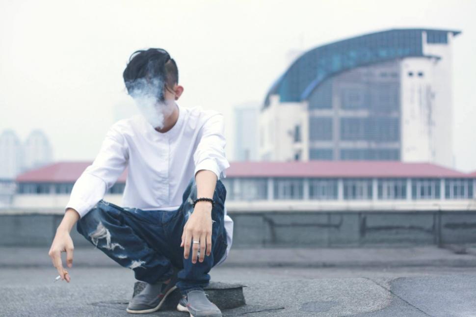 Free Image of Man squatting with smoke in front of building 