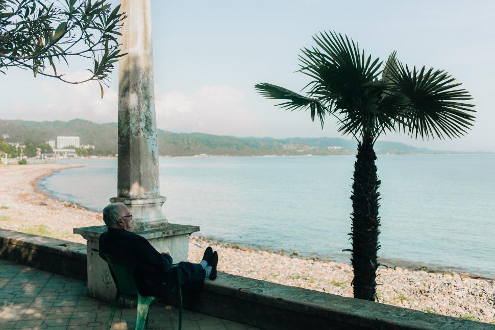 Free Image of Elder man resting by the sea under a palm 