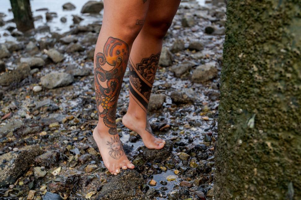 Free Image of Bare feet with detailed leg tattoos on rocky shore 