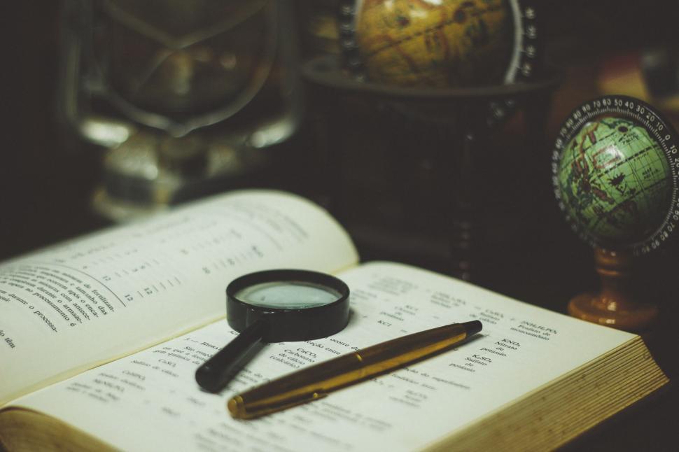 Free Image of Magnifying glass on open book with pen and globes around. 