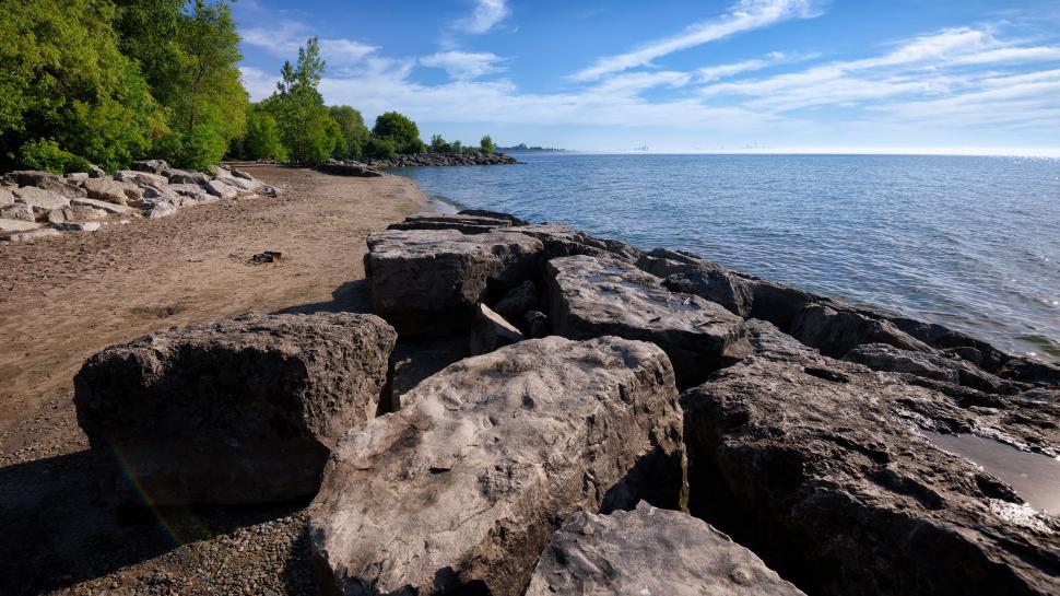 Free Image of Rocky lakeshore with green trees under blue cloudy sky 