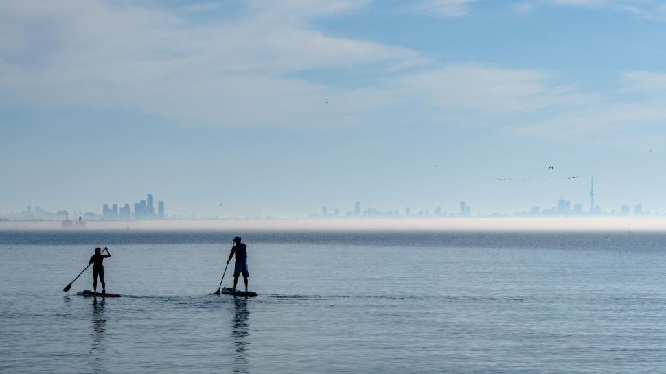 Free Image of Two people paddleboarding on tranquil water with cityscape view. 