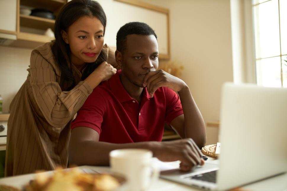 Concentrated young adult husband and wife looking at screen of modern laptop, reading article, surfing real estate or traveling websites, woman leaning on man s back, sitting at table in kitchen