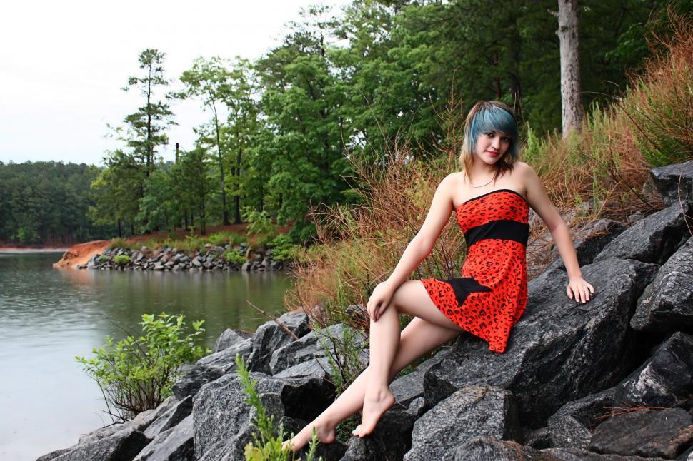 Free Image of A beautiful young woman posing in a red dress on rocks by a lake 
