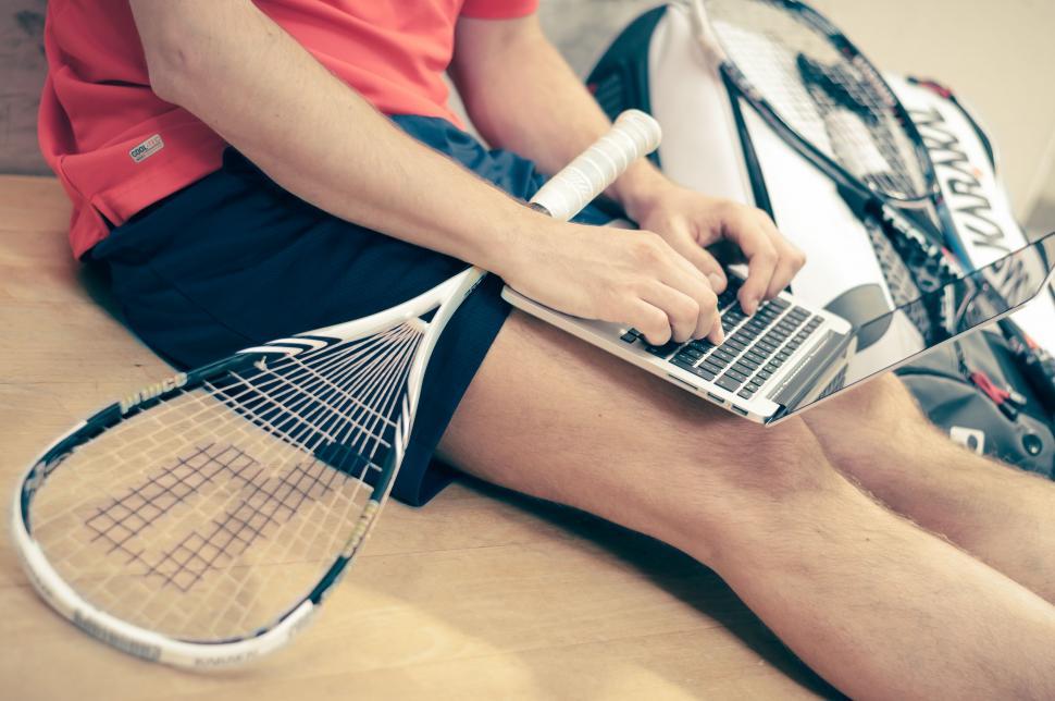Free Image of Man Sitting on Bed With Laptop and Tennis Racket 