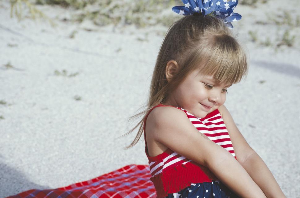 Free Image of Little Girl Sitting in Red, White, and Blue Dress on Sand 