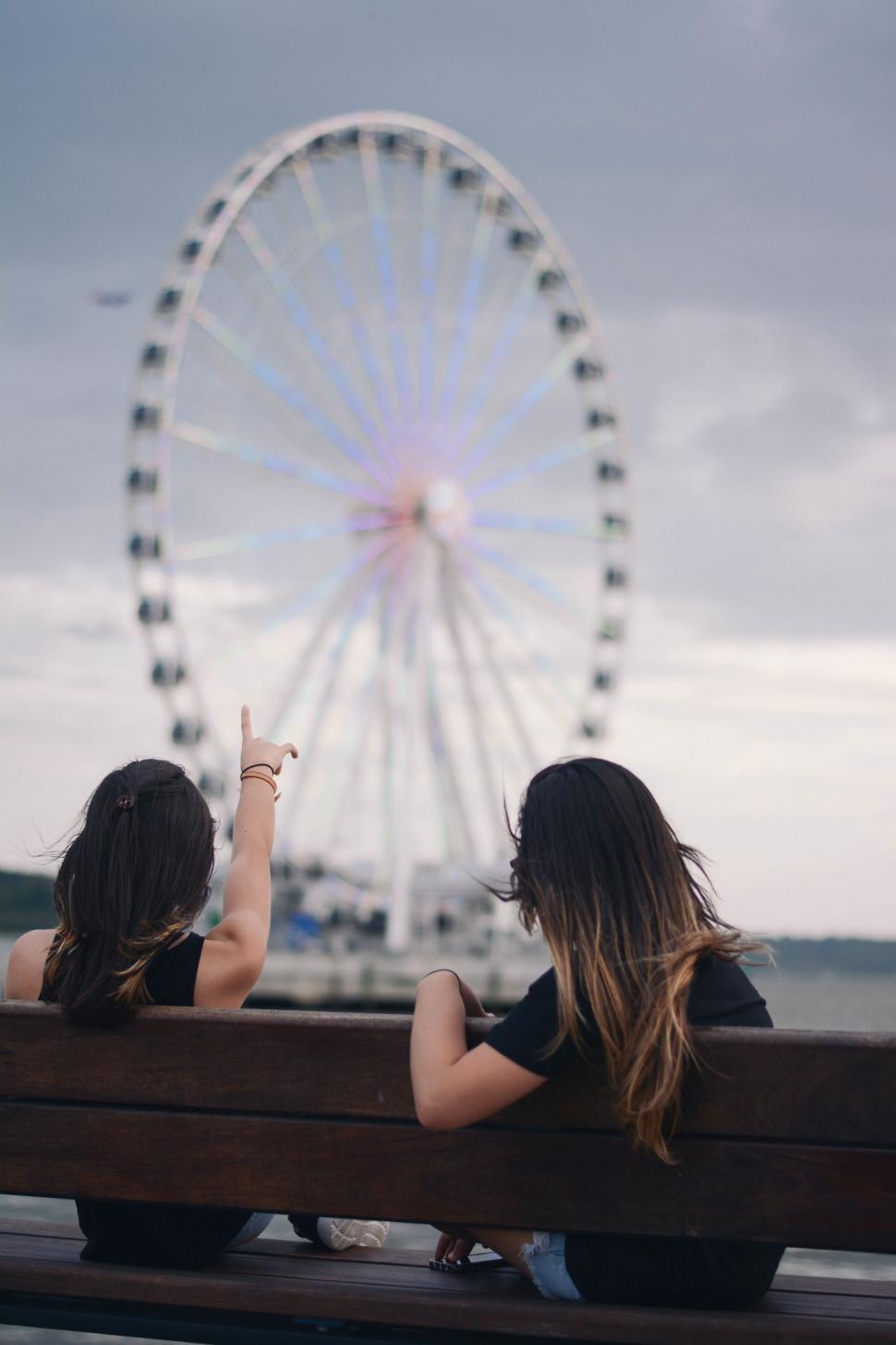 Free Image of Two Women Sitting on a Bench in Front of a Ferris Wheel 