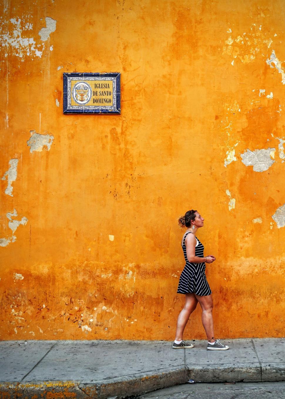 Free Image of Woman Walking Down the Street in Front of a Yellow Wall 
