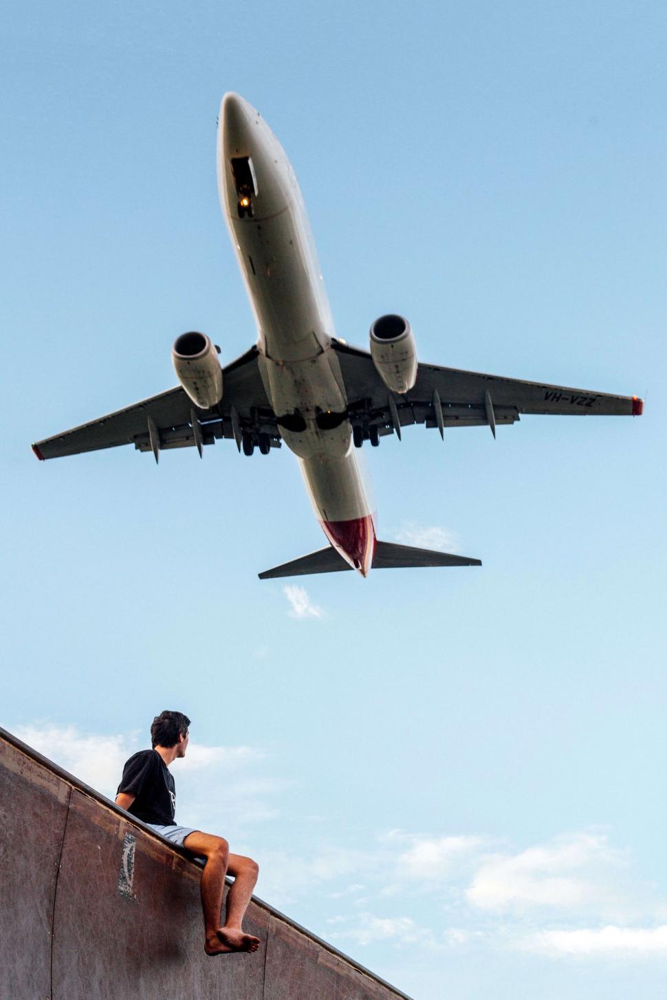 Free Image of Man Sitting on Ledge Watching Airplane Flying Overhead 
